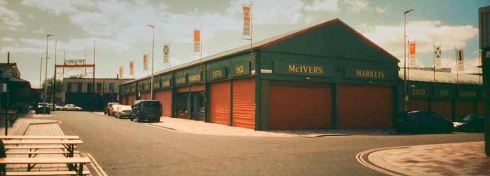 A colour panoramic photograph taken on 35mm film showing a junction in the Barras Market in Glasgow. There are benches to the left of frame and on the opposite corner one of the distinctive buildings that houses some of the extensive market. It is not a market day and the red shutters are closed. The Barras metal arch sign is in the distance further down the road. The signs on the market buildings say "McIver's Markets" in yellow painted on letters on a green backing. The sun is out and there are white clouds in the sky.