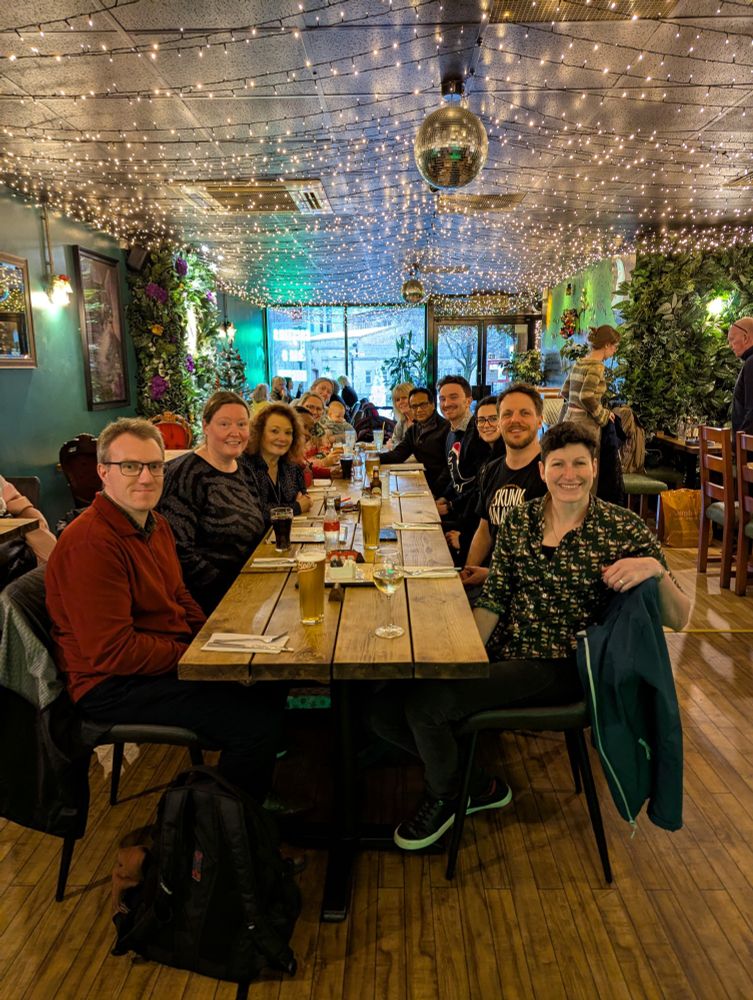 The NIHR Policy Research Unit in Behavioural and Social Sciences enjoying their festive celebrations last week. There is 12 people sat around a long wooden table on a dark wooden floor. There are drinks on the table, with silver cutlery on white napkins on the table. The walls are green and the ceiling has a combination of fairy lights and mirrored disco balls.