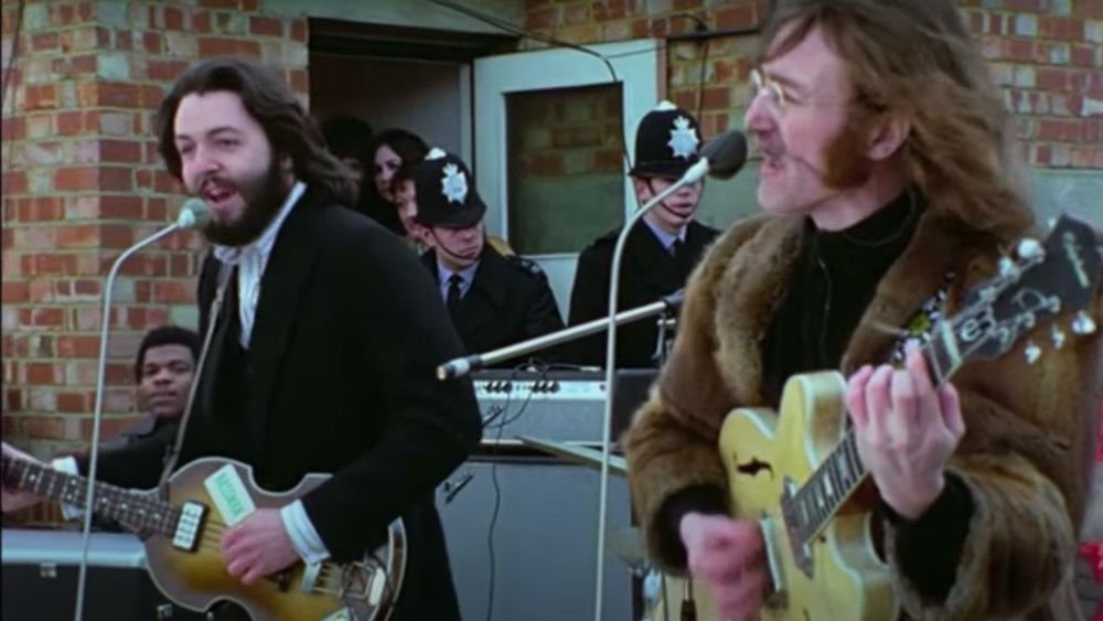 Paul McCartney and John Lenon preforming on a roof with several police officers wearing silly hats standing behind them. 