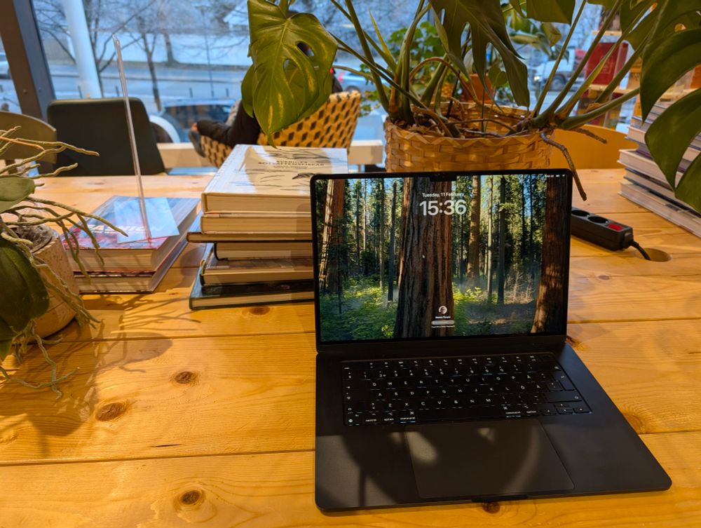 A black MacBook laptop is open on a wooden table, displaying a lock screen with a forest wallpaper and the time "15:36" on Tuesday, 11 February. The table has a natural, rustic appearance with visible knots in the wood. Surrounding the laptop are stacks of books, potted plants, and a power strip. The scene is set in a well-lit, cozy space with large windows in the background, showing an outdoor winter landscape with bare trees and a snowy ground. A person is sitting in a woven chair near the window, partially visible in the background.