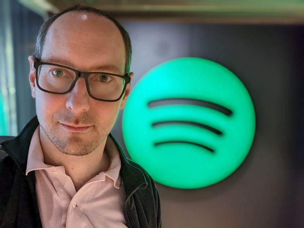 A selfie picture of Marcin standing in front of a large green Spotify logo in the "Urban Escape" office in central Stockholm.