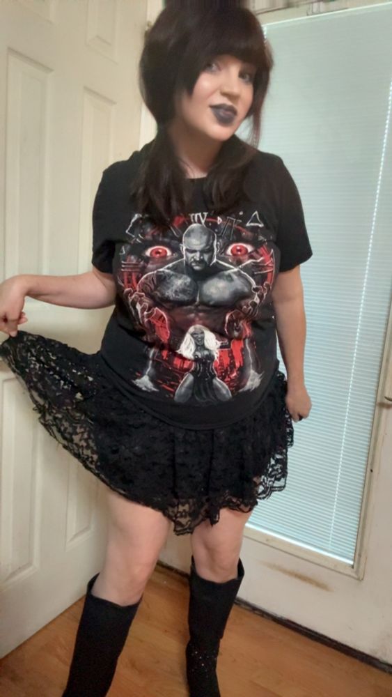 Serra stands on a wooden floor between two white doors. She wears knee high black boots, a lacy black skirt she holds to the side, and a Karrion Kross and Scarlett T shirt. Her natural hair is in pig tails and she wears black lipstick.