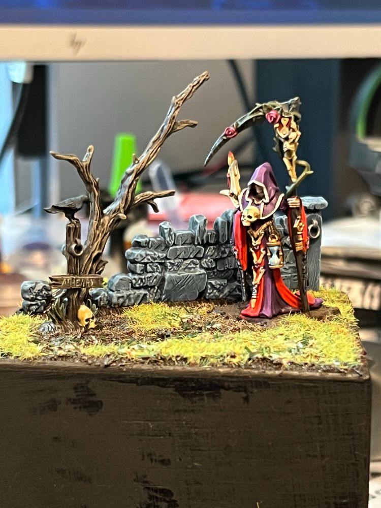 Necromancer in purple and red robes with a scythe. The scythe has vines with roses on it. Behind her is a crumbling wall and dead tree. Next to her is a sign reading “Athenry” with a crow atop it. The ground is patchy grass and earth. 