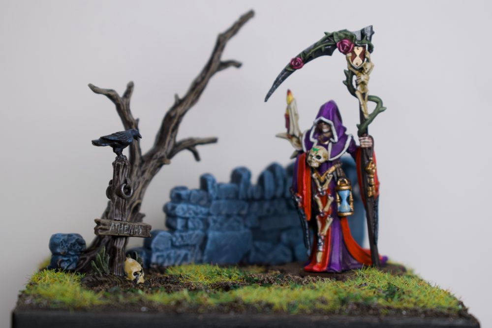 A diorama with a warhammer necromancer model, a crumbling wall and dead tree behind her, and a crow on a sign next to her. 