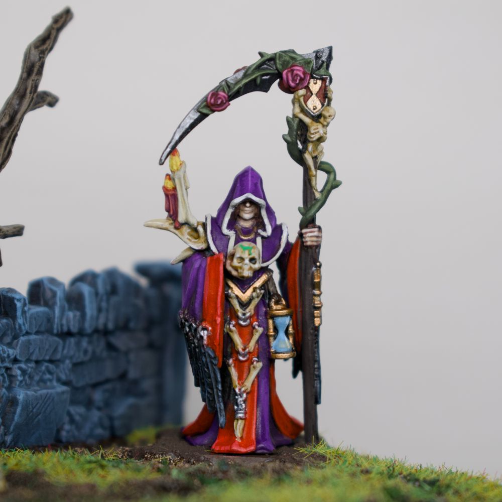 A closeup of the necromancer model. She has an hourglass, holds a skull, and in her other hand is a scythe with vines and roses on it. She has crow feathers on her sleeves and a crow skull on one shoulder. 