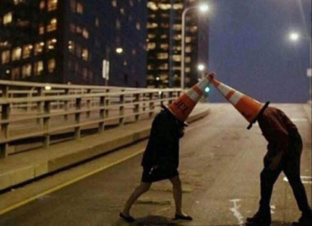 Two people standing on a bridge ramp, wearing traffic cones, and butting each other.