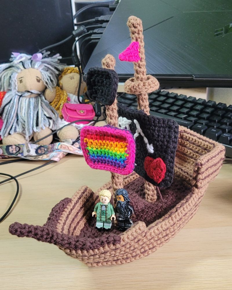 A photo of a crocheted pirate ship with Lego Ed and Stede standing on the deck. The front flag is a rainbow flag and the main flag is Blackbeard's flag. She is so cute!