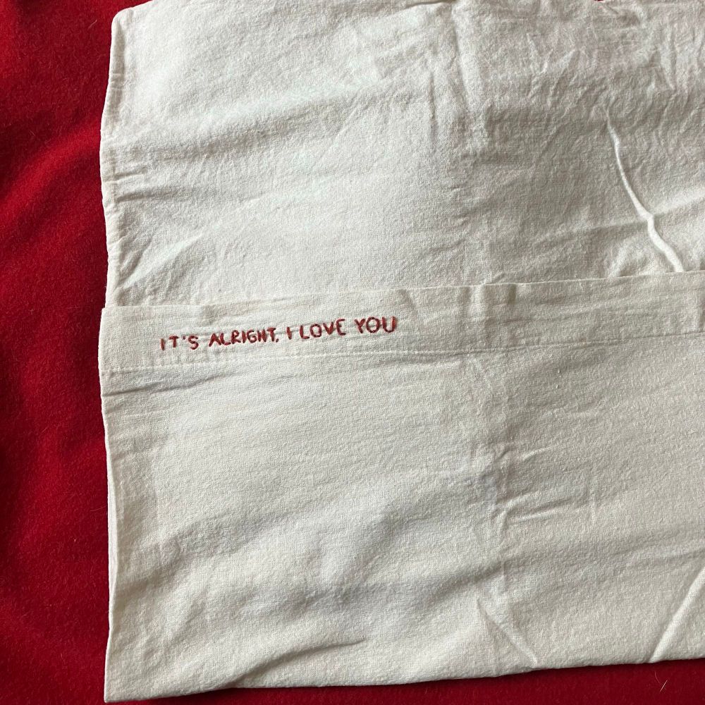 Photograph of a white pillowcase embroidered with the words ‘It’s alright I love you’.
