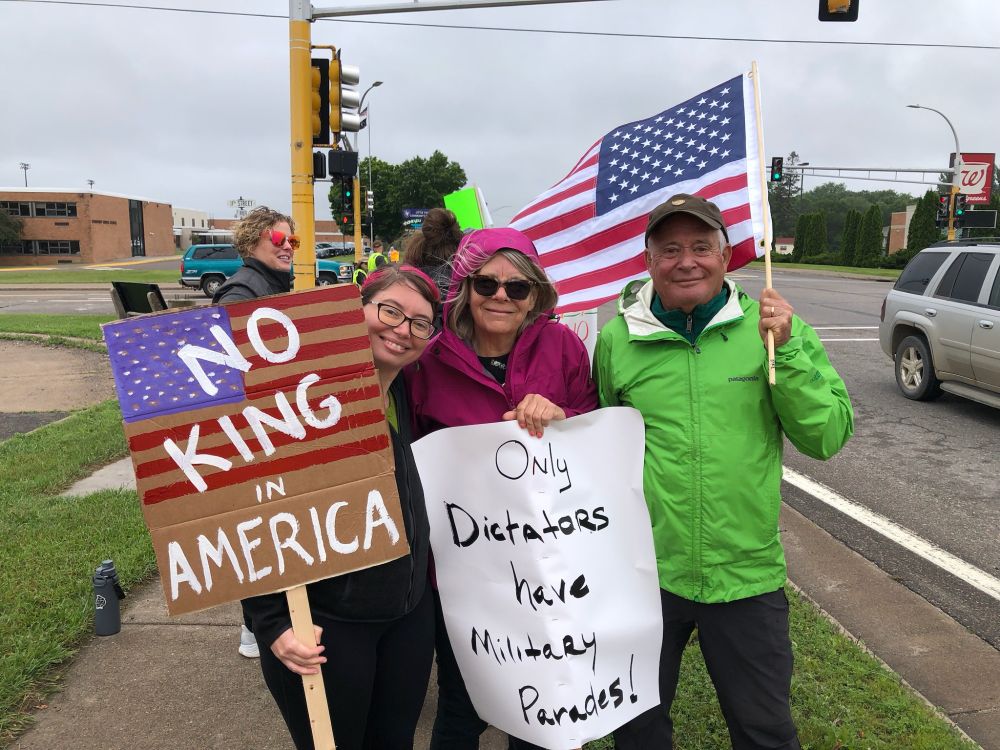 Protestors with an American flag and signs that say "No king in America" and "Only dictators have military parades"