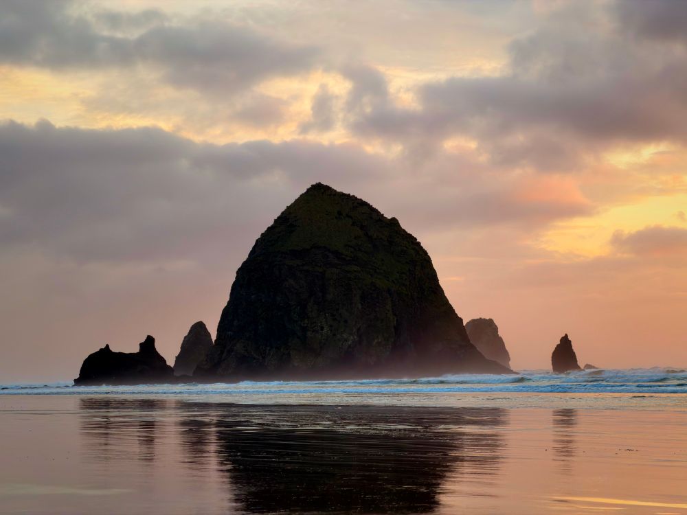 A profile of Haystack Rock. It’s reflecting off the wet sand. Waves are breaking at its base, creating some foreground mist. 