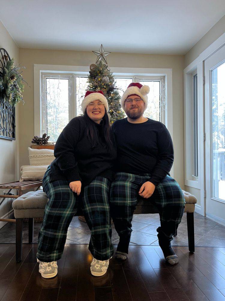a photo of rach and their partner tony, sitting on a bench in front of a christmas tree. both rach and tony are wearing santa hats and matching dark green plaid pyjamas with black sweaters