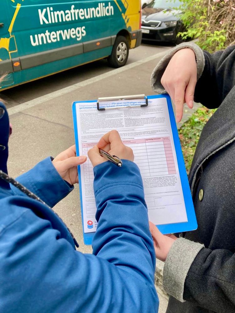 Zwei Personen stehen auf dem Trottoir. Eine Person unterschreibt den Unterschriftenbogen der Finanzplatz-Initiative auf einem Klemmbrett, das von der anderen Person gehalten und gezeigt wird. Im Hintergrund steht ein Lieferwagen mit der Aufschrift ‘Klimafreundlich unterwegs’.