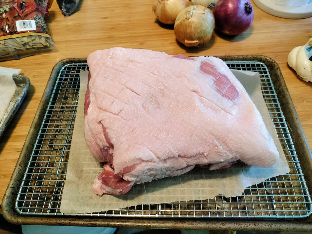pork shoulder with scored fat and salt crust on a tray. onions in the background