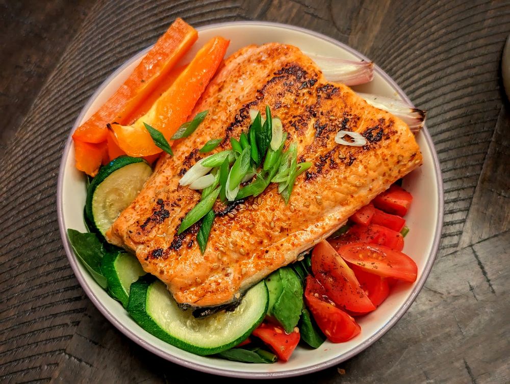 A picture of a piece of blackened salmon sitting on top of a salad with roast veggies and sliced tomatoes.  It looks tasty, mostly because it was.