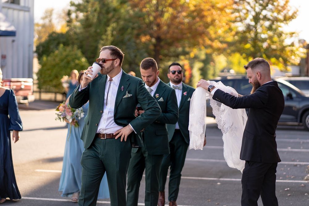 In the foreground, a statuesque man in brown sunglasses and green suit is drinking from a white paper cup filled with espresso and hot water. In the background are members of the wedding party including two other men in green suits, several women in blue dresses, and the officiant in a black suit holding the veil.