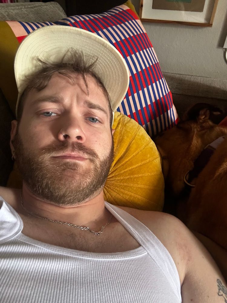 Man in white tank top and baseball cap laying on couch (sweaty) with dog.