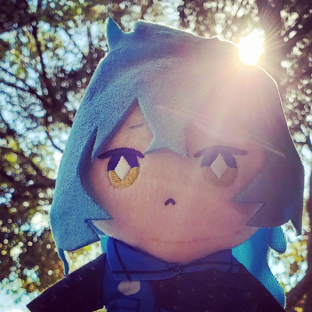 Idia Shroud Nui plush back-lit by the sun. There are lovely trees around, as well as a nice blue sky!