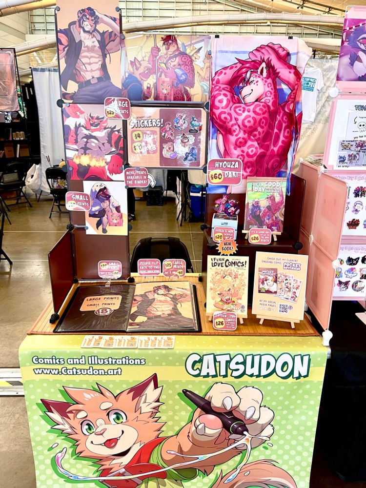 A photo of my dealers den booth. It is a half size table featuring my books, prints, stickers and acrylic standees.