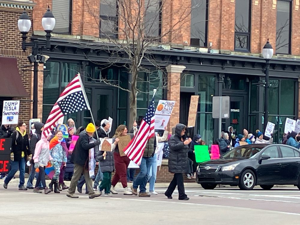 Protestors carrying signs and American flags (one upside down) crossing the street as others line the sidewalks.