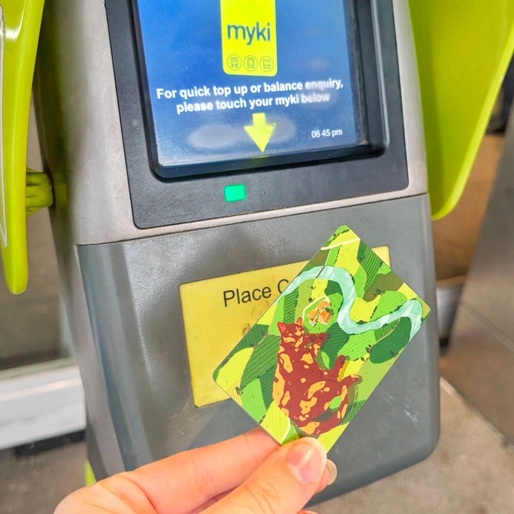 Photo of me checking the balance on my Myki travel card. The card is decorated with my travel card vinyl sticker, showing a giant tortoiseshell cat sleeping peacefully on a big slice of farmland, with a river going by it.