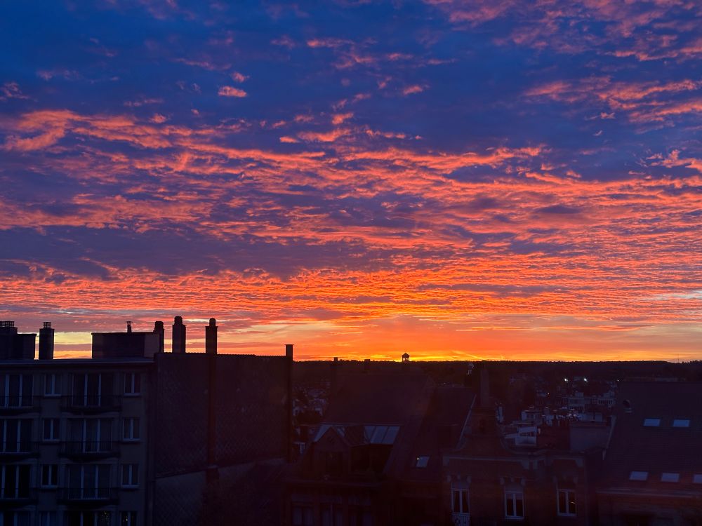 Cloudy sunrise. Clouds are orange and red. Below, the city buildings are almost black. This is a view of a part of Brussels.