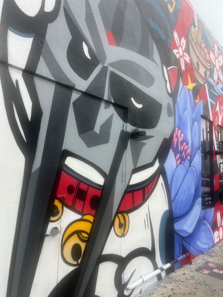 Image of a mural with MF DOOM’s distinctive face mask on a lucky cat, with a graphic flower motif to the right. At the edge of the photo is what appears to be a storefront.