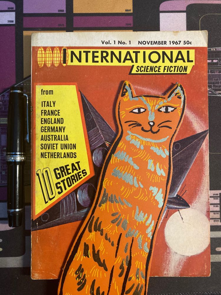 Photo of the first issue of International Science Fiction with my tabby cat bookmark and fountain pen. Cover (mostly behind the bookmark, sorry) shows a spaceship with a bunch of flags. 