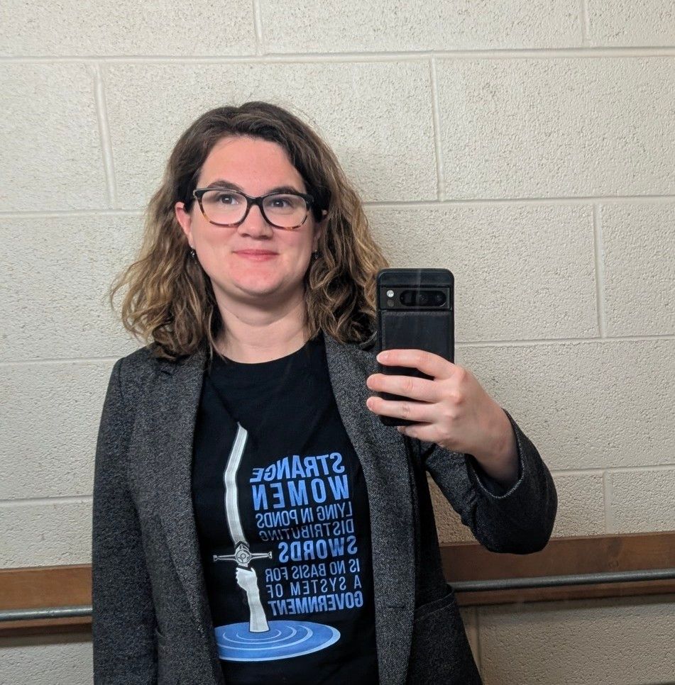 A bathroom selfie of me wearing a grey blazer and a t-shirt that says "Strange women lying in ponds distributing swords is no basis for a system of government" and a graphic of the hand of the Lady of the Lake holding Excalibur aloft.