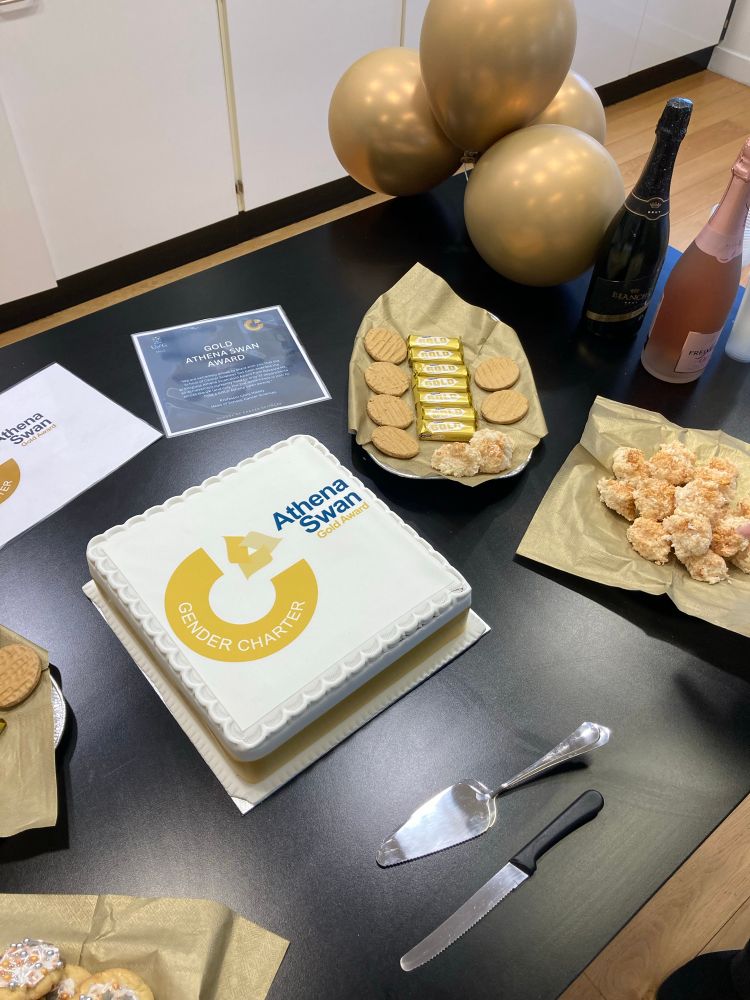 An Athena Swan Gold Award printed cake, ornately presented on a table with various other gold-themed cakes, bottles of sparkling wine, gold balloons & napkins, a cake knife and a server.