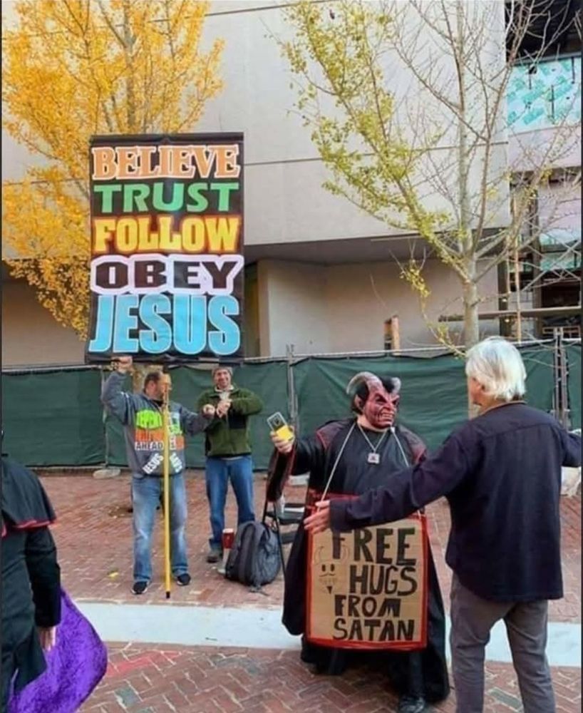 2 men holding a sign that reads: BELIEVE, TRUST, FOLLOW, OBEY JESUS. Next to them is a man in a long black coat and goat horns wears a sign around his neck that reads: Free hugs from Satan, a man with his arms outstretched is walking towards him.