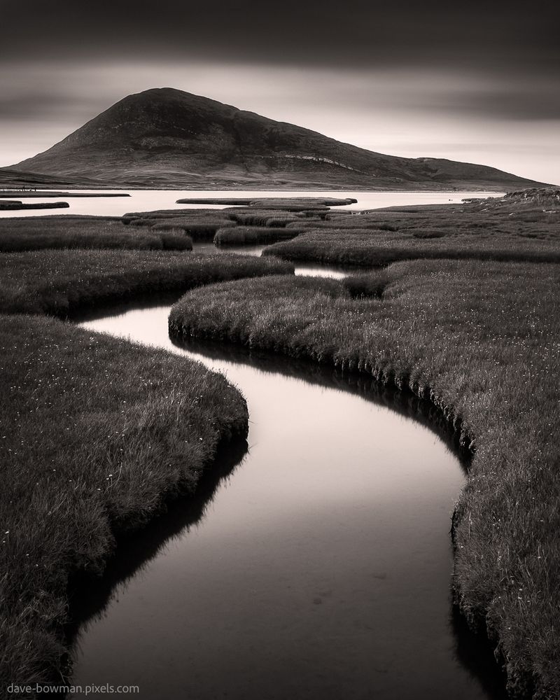 In this monochrome photograph captured on the Isle of Harris in the Outer Hebrides of Scotland, the Northton Saltmarsh takes centre stage. With its mesmerising beauty bathed in a subtle warm tone, the image exudes a sense of tranquility and serenity. A long exposure lends a dreamlike quality to the scene as the saltmarsh channels wind their way towards a distant mountain. Above, the ethereal clouds shift gracefully across the sky, blurring softly, adding to the otherworldly ambiance. This arresting image invites you to explore the mystical allure of a remote and untouched landscape, where time seems to stand still.