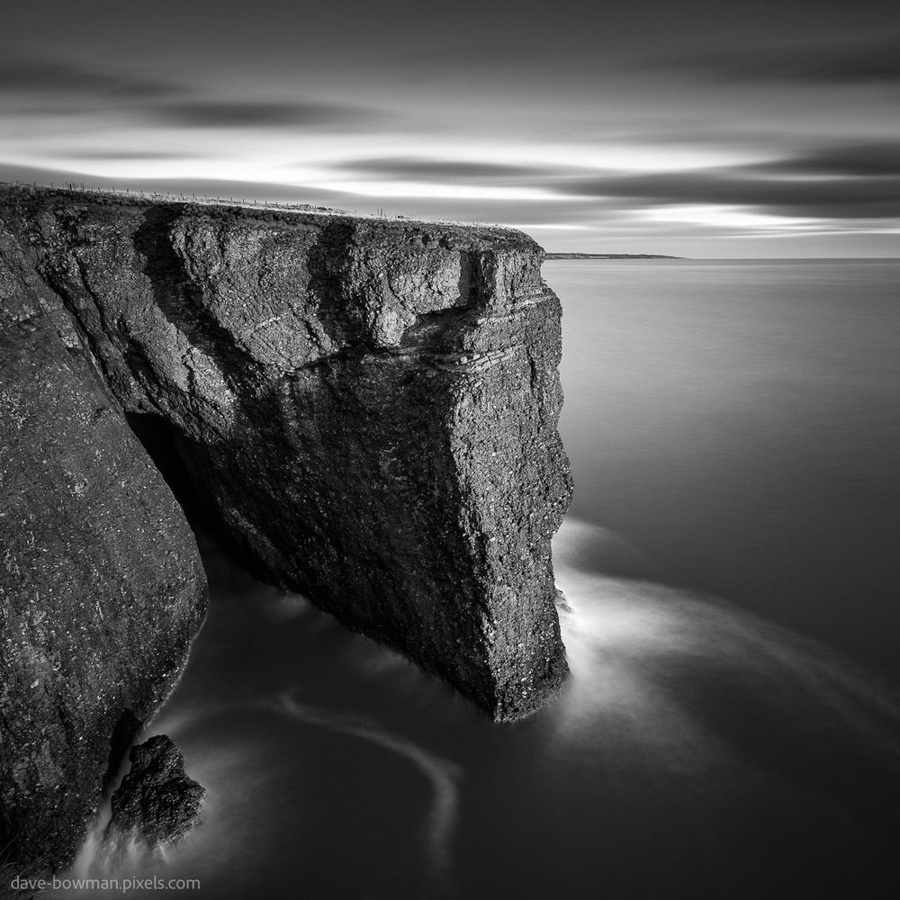 A photograph of a towering cliff face rising sharply from the edge of the sea on the Scottish coastline. The rock surface is rugged and craggy, showing deep textures and jagged edges that emphasise its dramatic height. At its base, the ocean appears smooth and mist-like, blurring into a soft, silken surface that contrasts with the harshness of the stone. The scene is presented in strong contrasts of light and dark, which draw attention to the power and raw beauty of the cliffs against the calm expanse of water below.
