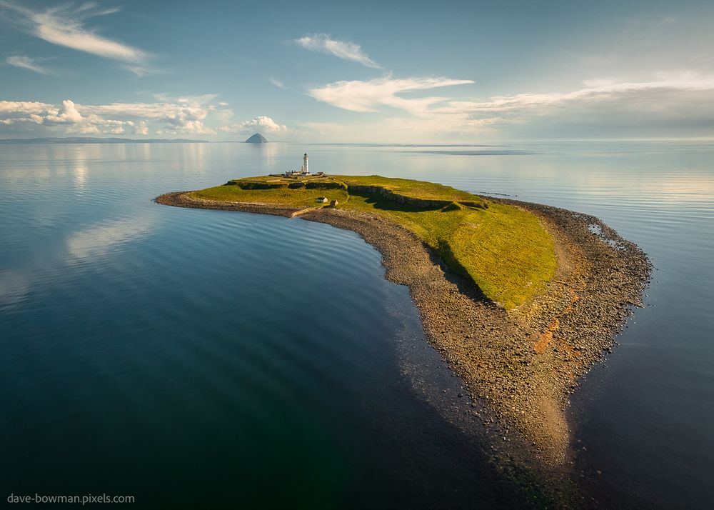 A photograph of Pladda Island taken from above, showing the full shape of the island surrounded by calm blue sea. Near the far end of the island, the small but clearly visible Pladda Lighthouse sits close to the shore. The land is mostly green with patches of exposed rock. In the distance across the water, the distinct dome of Ailsa Craig rises on the horizon beneath a clear blue sky.