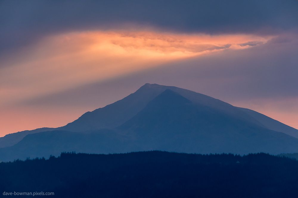 A photograph of a dramatic mountain landscape in Snowdonia National Park (Eryri), North Wales. At the centre, a dark mountain stands in silhouette beneath a sky where warm rays of sunlight pierce through a thick layer of cloud. In the foreground and middle distance, a series of softly contoured hills overlap and fade into the distance, each one darker than the last, creating a sense of depth. Behind it all, the sky glows with the soft, warm hues of twilight, adding contrast and atmosphere to the scene.