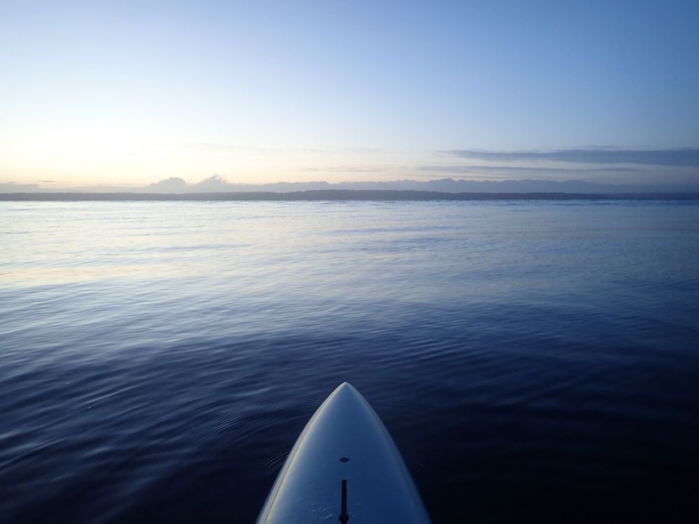 The pointed nose of a pale-blue prone paddleboard points across the glassy water of the Puget Sound toward the distant Olympic Mountains. The sky is clear other than a few low clouds, and the fading light of twilight is reflected in the water and board.
