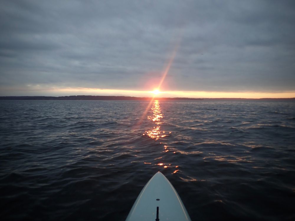 The sun appears on the horizon, wedged between gray clouds above and the dark waters of Puget Sound below. The sun's bright red light is reflected off the water. The pointed nose of a pale-blue prone paddleboard sits in the foreground, where the paddler has stopped to take the photo.