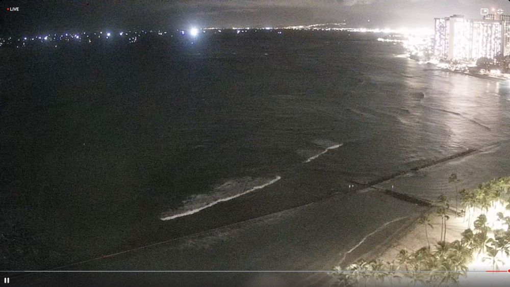 A view from Waikiki beach, with the beach and city lights to the right and the ocean to the left. In the distance at sea are dozens of lights along the horizon where boats have evacuated to wait out the tsunami in deep water.