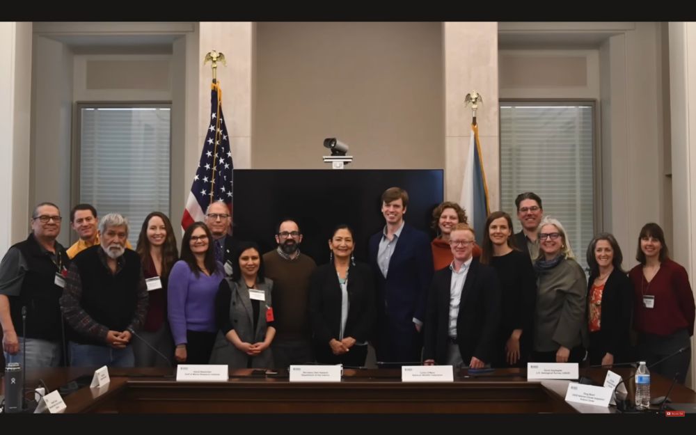 Members of the Advisory Council for Climate Adaptation Science stand with former US Department of the Interior Secretary Deb Haaland at center. The group of 17 is smiling, standing behind a dark table in a white room with flags, windows and a large screen at their back.