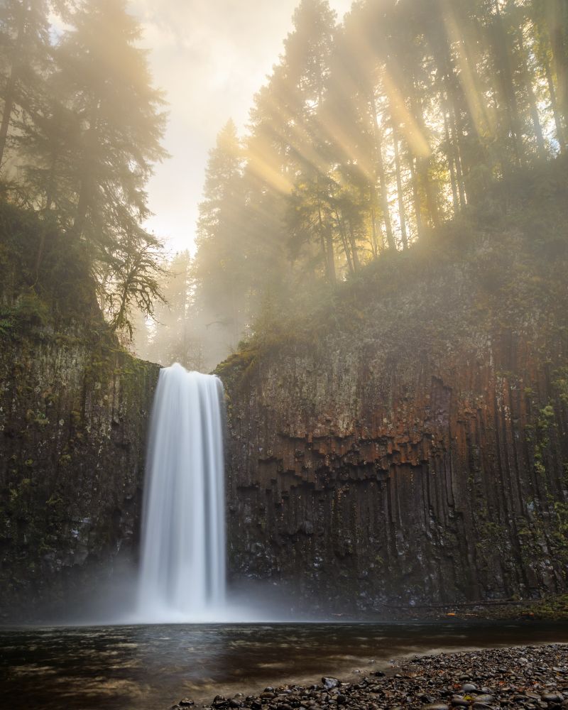 Silky smooth waterfall coming down a reddish basalt cliff with yellow sun rays coming through the trees on the top of the cliff