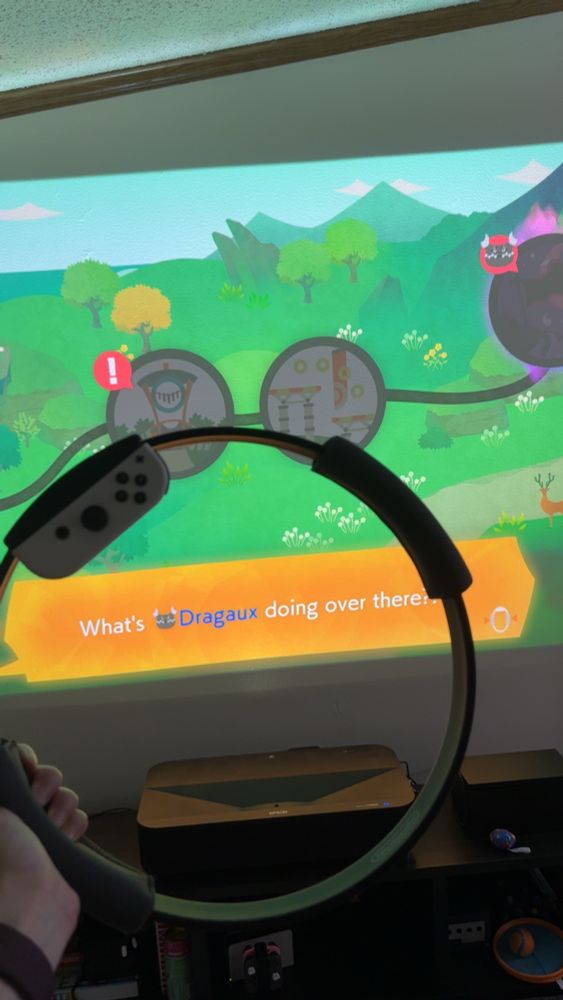 A hand is holding the ring from Nintendos Ring Fit Adventure, the game’s menu screen is on the tv in the background 