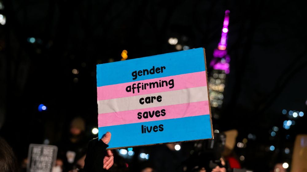 A close-up of a sign held at a nighttime protest. The sign has the colors of the transgender flag (blue, pink, and white) and reads, "Gender affirming care saves lives." The background shows blurred lights and the outline of a brightly lit tower in purple.