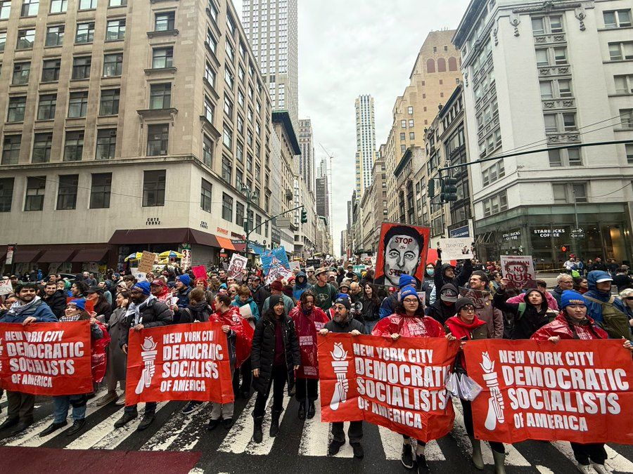  A large crowd of protesters marches down a city street lined with tall buildings. At the front, people hold four bright red banners that read "New York City Democratic Socialists of America" with a white torch logo. Many participants wear red ponchos or winter clothing. Behind them, others hold signs including one that says “Stop the Genocide,” another with a face illustration, and others expressing political messages. The protest takes place in Midtown Manhattan, with recognizable buildings and a Starbucks storefront visible.