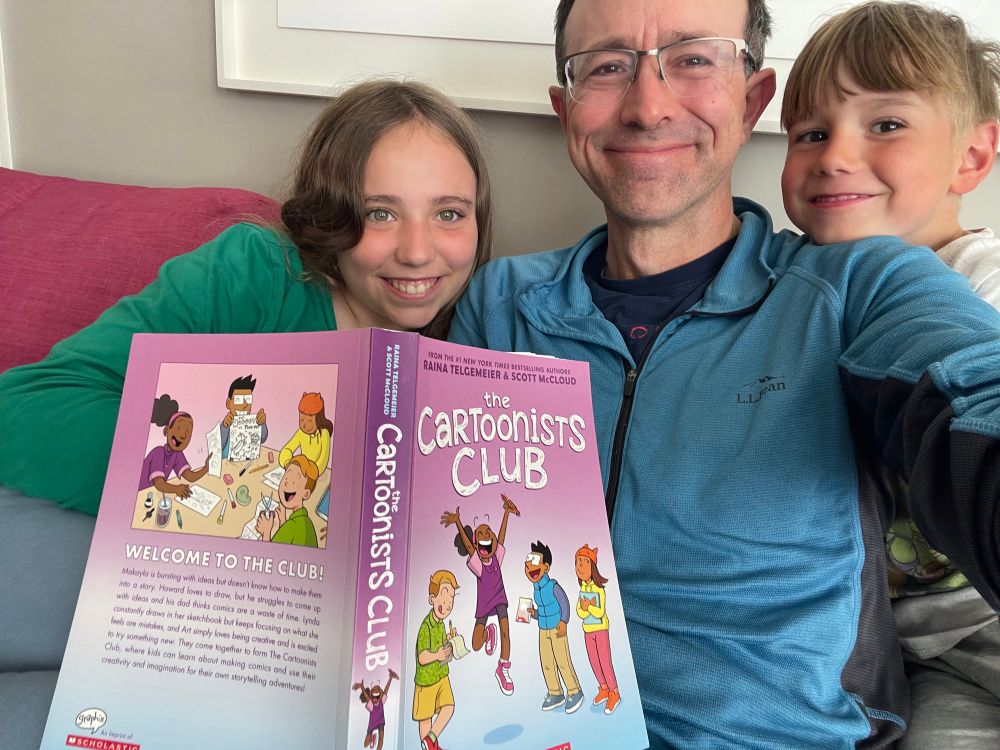 Selfie of me holding up the cartoonists club book with big kiddo and little kiddo all looking at camera smiling and being generally silly 