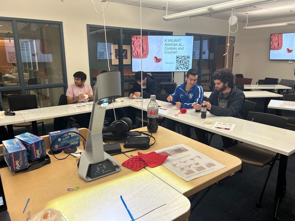 A group of students learning crochet. A slide reads “Cookies and Crochet for VAL-entine’s Day”