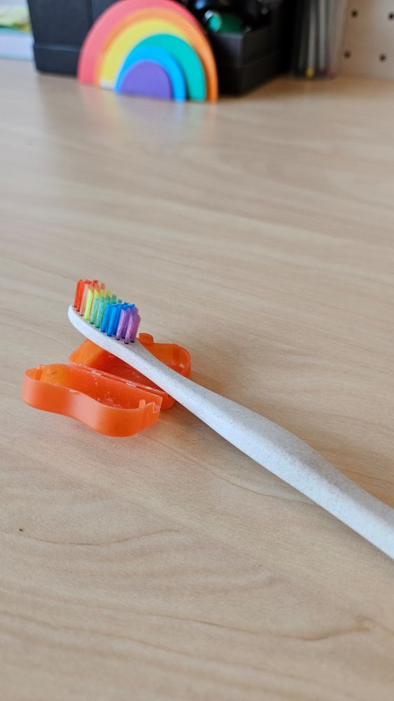 Ein tannbørste med regnboge-bust liggende på jobbpulten min. I bakgrunnen synest ein post-it-regnboge mot pennehaldar-systemet, som er svarte boksmodular.