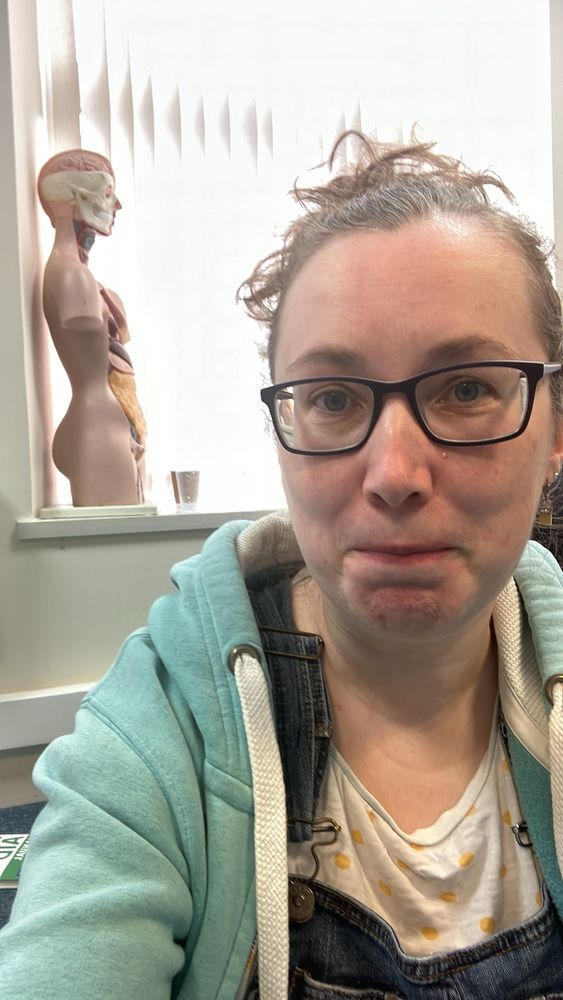 A white woman with glasses sitting on floor with a model showing body organs behind her on a windowsill. There’s a first aid manual on floor behind her too.