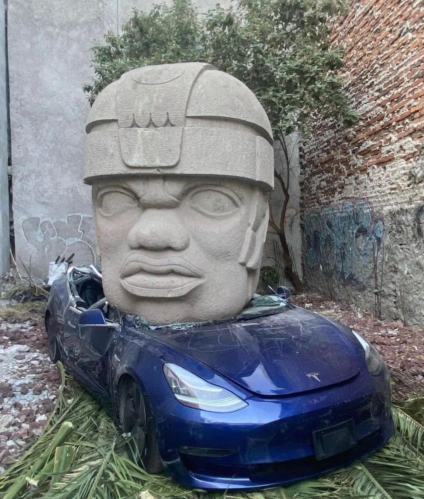 At the Mexican CMDX, a concrete replica of an Olmec head fell off the crane and crushed a Tesla parked in an alley. The Olmec head, which is about 7' tall is facing the camera; the crushed Telsa is a metallic deep blue, and there's lots of broken safety glass. It's on palm fronts and some gravel, with a brick wall in the background to the right. Behind it, there's some obscured graffiti in Spanish.