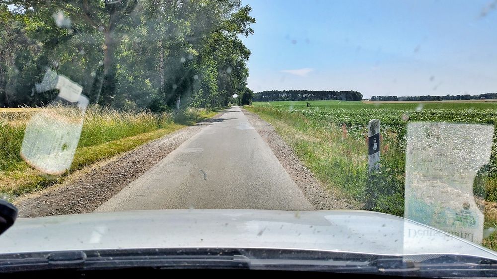 Landstraße mit Straßenschäden links und rechts der Fahrbahn, sodass zwei Fahrzeuge nur schlecht aneinander vorbei kommen. Links Bäume, rechts Felder. 