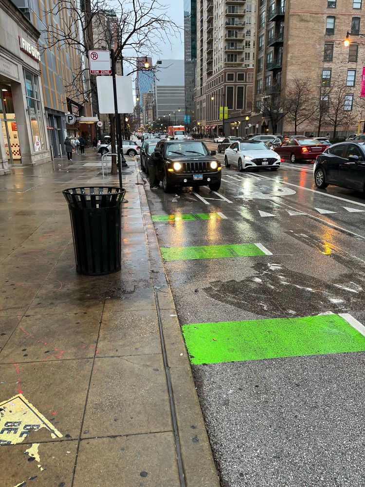 Three stripes of green paint on a southbound bike lane on Wabash, with three parked cars blocking the bike lane. The right turn lane for cars is left of the bike lane.