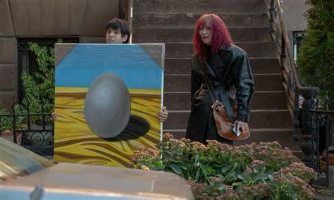 Screencap of Julio Torres and Tilda Swinton from the movie Problemista. Julio Torres is holding a large canvas: a painting of an egg on yellow silk with a blue background. 